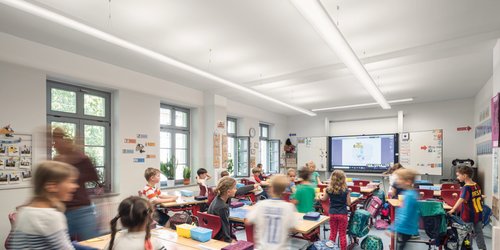 Grundschule Ein großes längliches Klassenzimmer mit parallel zum Fenster angebrachten Pendelleuchten, die von der Tafel bis an die gegenüberliegende Wand reichen und viel Helligkeit geben.